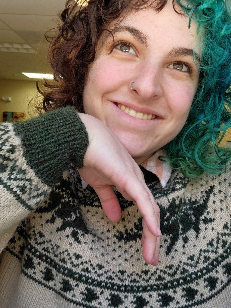 Casey wearing her sweater. She is looking up into the distance and resting her head on her hand. You can see the green stegosauruses on the yoke of the sweater, and the green colorwork detailing on the cuff. 